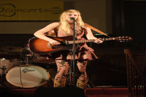 Eine Frau, die auf der Bühne Gitarre spielt und singt, mit einer Band im Hintergrund und einem Bildschirm an der Wand.