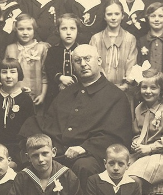 Ein Schwarz-Wei├č-Foto eines ernst aussehenden Mannes, der in der Mitte sitzt und von einer Gruppe Kinder umgeben ist, die bewundernd zu ihm aufschauen.