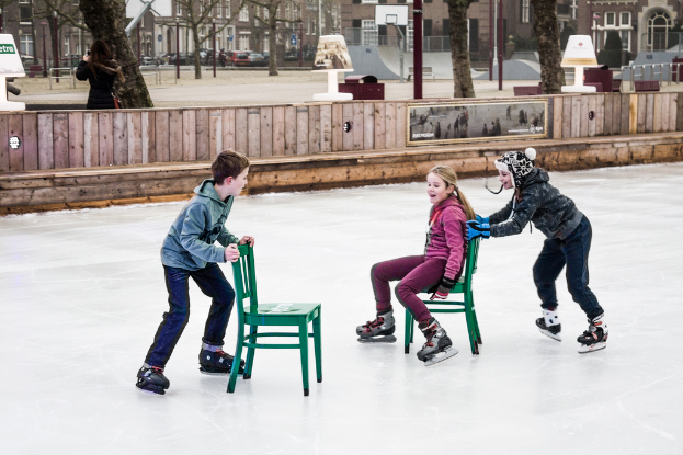 Kinder beim Skifahren vor einer Spielplatzkulisse mit drei Kindern und zwei Stühlen in der Mitte und Gebäuden, Bäumen, Bänken, Pfosten und einem Basketballfeld im Hintergrund.