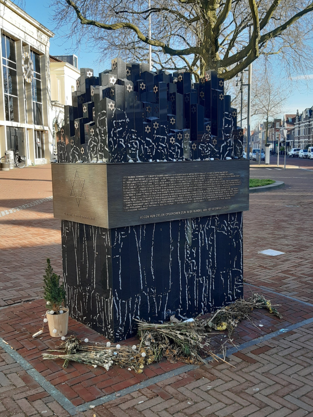 Denkmal in einem Stadtplatz für Holocaust-Opfer, mit einem Baum davor, einem Topfgewächs auf dem Boden, verstreuten trockenen Blättern und Hintergrundgebäuden, Fahrzeugen, Pfählen und Bäumen.