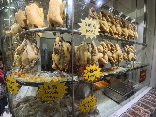 Vitrine mit ganzen gerösteten Hühnern auf einer Theke, eine Person steht links daneben, Deckenlampen im Hintergrund und Tabletts mit Lebensmitteln auf der rechten Seite.