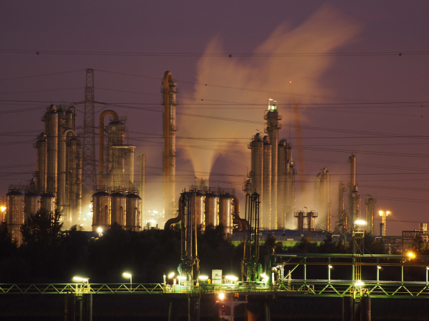 Ölraffinerie bei Nacht mit Rauch, der aus ihr aufsteigt, beleuchtet von umliegenden Lichtern, vor einem sichtbaren Himmel, mit einem Wasserzeichen.
