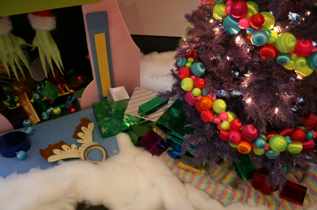 Drinnen steht rechts ein geschmückter Weihnachtsbaum mit Lichterkette und Schmuck, in der Mitte Geschenkboxen und Dekorationen, in der oberen linken Ecke eine Hütte mit Dekorationen und unten Wolle und Watte.