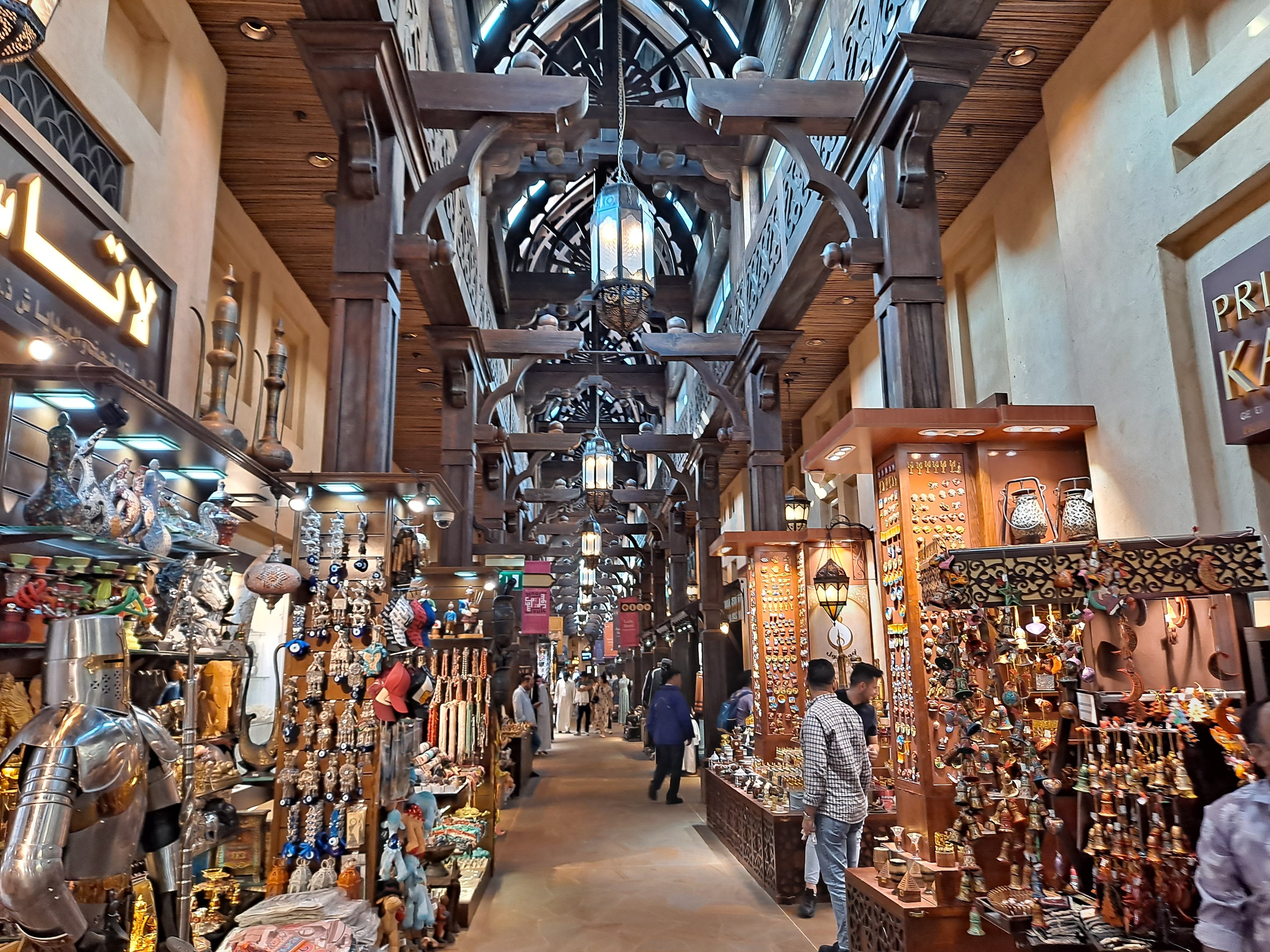 Ein geschäftiges Dubai-Markt mit Menschen, die zwischen Ständern mit Gegenständen hindurchgehen, Wänden mit Texttafeln und einer Decke mit warmem Licht.