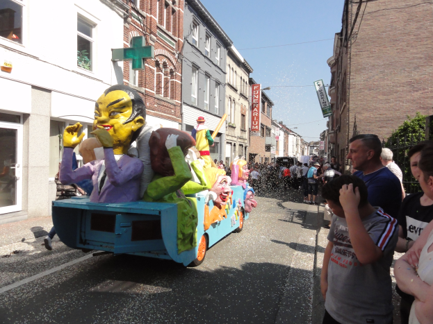 Eine Gruppe von Menschen auf einem bunt verzierten Spielzeug-Schwimmwagen bei einem Umzug, mit Gebäuden, Texttafeln, Bäumen und einem klaren blauen Himmel im Hintergrund.