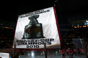 Eine nächtliche Stadionszene mit einem großen Banner in der Mitte, Zuschauern im Hintergrund, einem Glas auf dem Boden, einigen Spielern auf dem Feld und beleuchteten Tribünen.