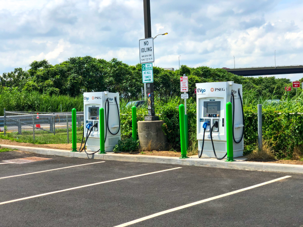 Eine Ladestation für Elektrofahrzeuge auf einem Parkplatz, umgeben von verschiedenen Geräten, Hinweistafeln, Vegetation und einer Brücke im Hintergrund bei bewölktem Himmel.
