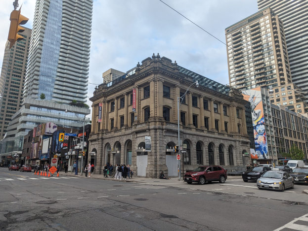 Eine belebte Stadtstraße mit hohen Gebäuden im Hintergrund, Fahrzeugen, Fußgängern, Verkehrskegeln, Laternen, Ampeln, Schildern, Werbetafeln, Bäumen und der Toronto Public Library als Mittelpunkt unter einem bewölkten Himmel.