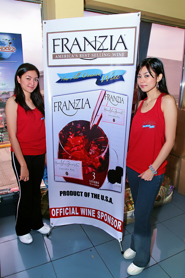 Zwei Frauen neben einem Plakat für Franzia, Amerikas bestverkaufendem Wein, mit einem Glasfenster im Hintergrund.