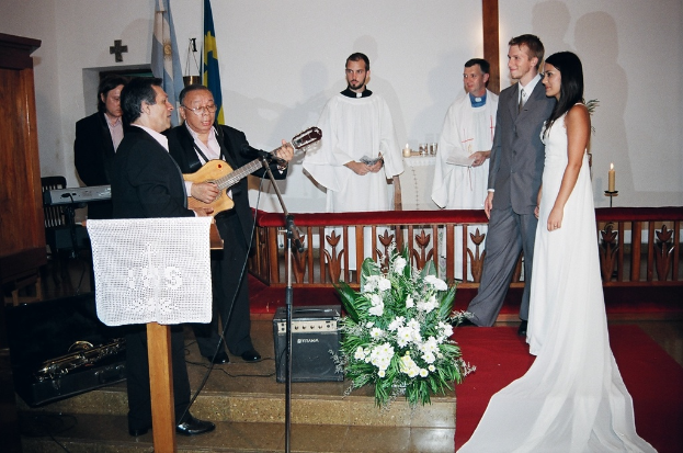 Eine HochzeitsSzene in einem Raum mit zwei Pastoren, einer Braut, einem Bräutigam und drei Musikern; ein Podium mit Stoff vor den Musikern, ein Tisch mit Kerzen hinter den Pastoren, ein Kerzenhalter hinter dem Paar und ein Bukett, Lautsprecher, Taschen und roter Teppich auf dem Boden; Flaggen, eine Wand mit einem Kreuz, einen Schrank und einen Stuhl im Hintergrund.