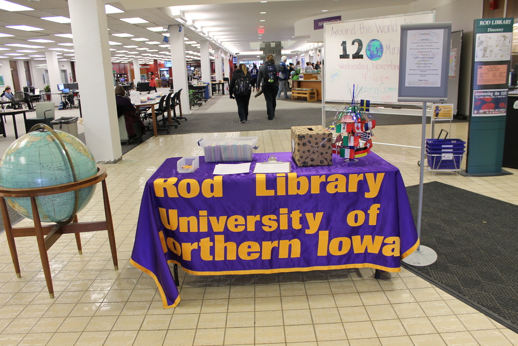 Eine Ausstellung mit einem Tisch, auf dem verschiedene Landesflaggen, eine Box, Papiere und ein Banner liegen, eine Kugel zur Linken, ein Anschlagbrett zur Rechten mit einigen Menschen in der N├Ąhe und mehreren Tischen und St├╝hlen zur Linken.
