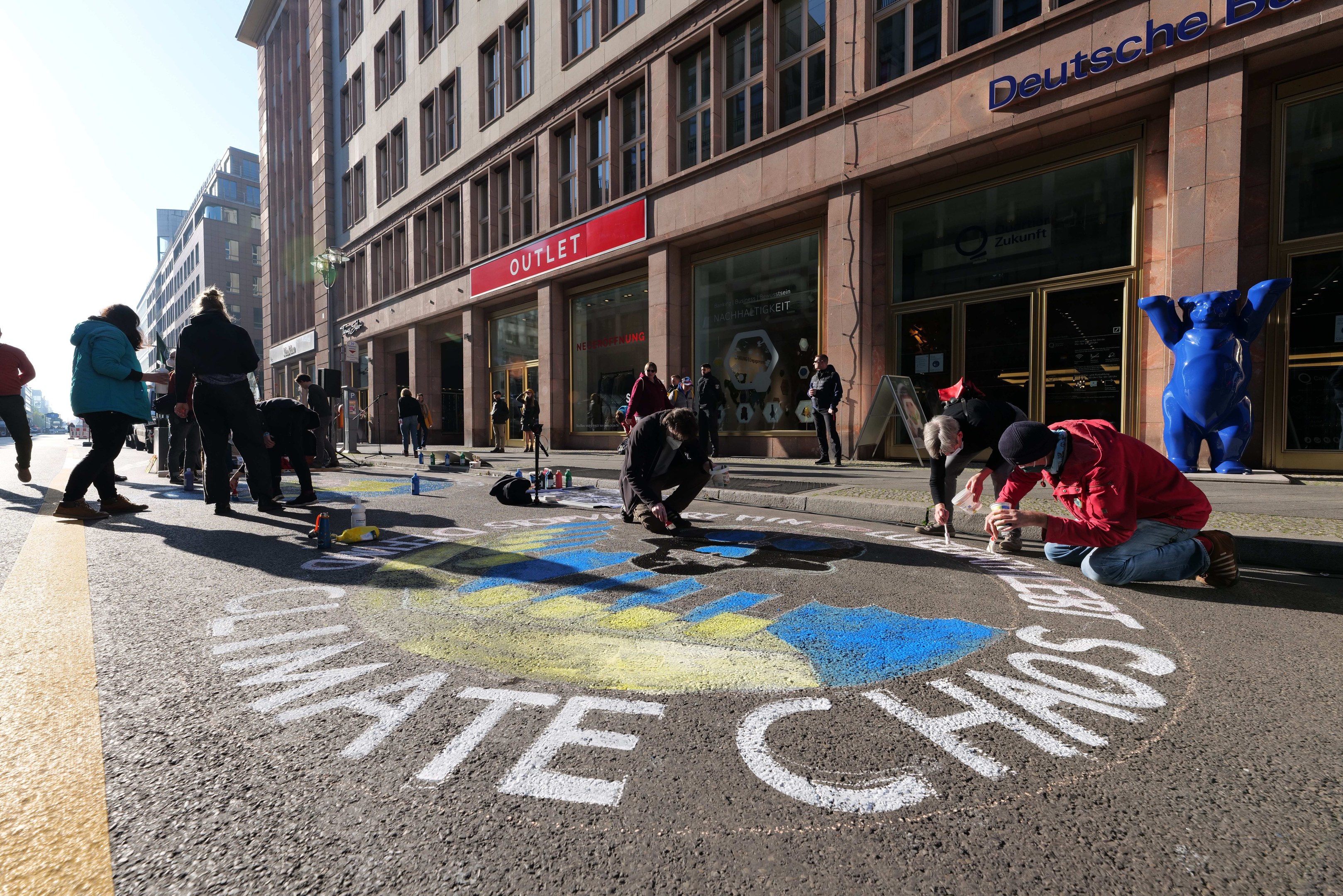 Gruppe von Menschen, die vor einem Gebäude mit Fenstern und Namensschildern sitzen, umgeben von Flaschen und anderen Gegenständen, mit Bäumen und einem klaren blauen Himmel im Hintergrund.