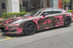 Ein Porsche Panamera mit Tarnfolie auf der Seite einer Straße geparkt, umgeben von Gebäuden, Bäumen, Pflanzen und anderen Gegenständen.