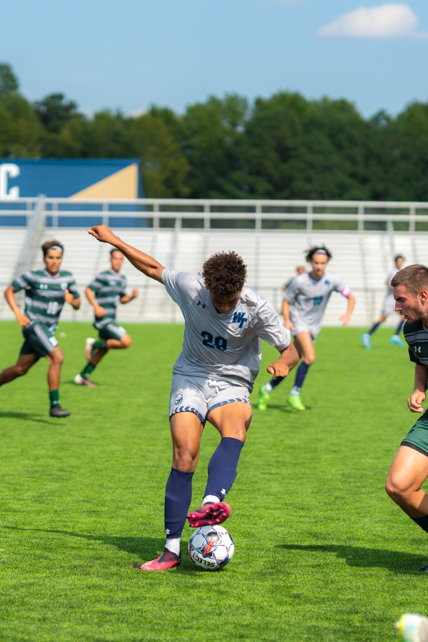 Eine Gruppe junger Männer, die Fußball auf einem Feld mit Bäumen im Hintergrund unter einem klaren blauen Himmel spielt, wobei ein Spieler Turnschuhe trägt.