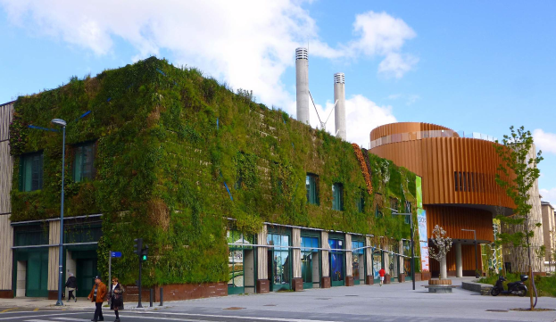 Ein große Gebäude mit einer grünen Wand an seiner Seite, umgeben von Straßeninfrastruktur, Fahrzeugen, Fussgängern, Bäumen und einem bewölktem Himmel, Teil des Green Building Project.