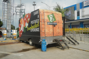 Lkw vor einem Gebäude mit einer Tafel, auf der "Teilen Sie ein positiv überladenes Erlebnis" steht, mit Stufen, einem Zylinder auf dem Boden, Geländern, Strommasten mit Drähten, Laternenpfählen, Bäumen und einem bewölkten Himmel.