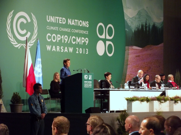 Eine Frau steht neben einem Podium mit Mikrofonen und spricht zu einer sitzenden Menge, mit einem Tisch voller Gegenstände, Flaggen, Pflanzen und einer weiteren Person in der Nähe.