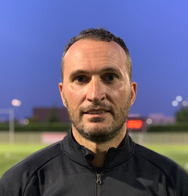 Ein Mann in einer schwarzen Jacke mit Logo steht auf einem Fußballfeld und sieht aus wie ein Trainer, mit einem unscharfen Hintergrund.