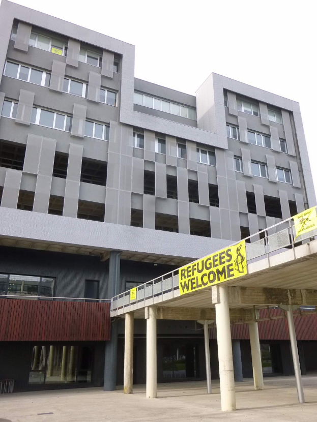 Ein großes Gebäude mit einem "Flüchtlinge Willkommen"-Schild, flankiert von Säulen und einem Geländer, vor einem Himmel als Hintergrund.