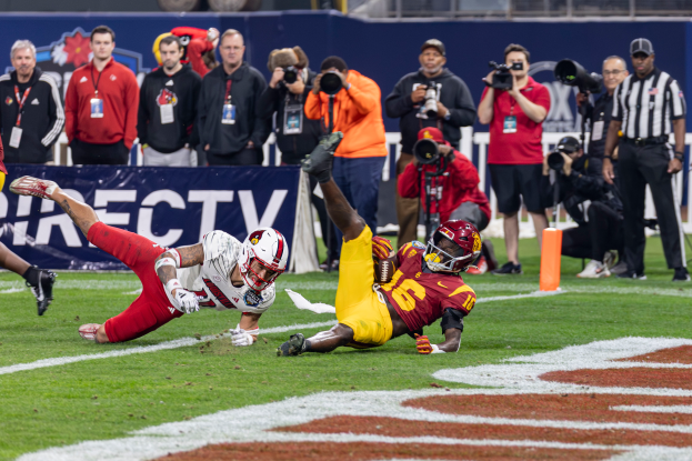 Ein Fussballspieler taucht in die Endzone für einen Touchdown, mit einer Gruppe von Menschen am Boden, einige halten Kameras, und ein Banner mit Text im Vordergrund, und Geländer, Pfosten und andere Gegenstände im Hintergrund.