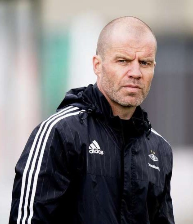 Ein kahlkopfiger Mann in einer schwarzen Jacke, der scheinbar ein Trainer ist, steht auf einem Fuballfeld und trägt eine marineblaue Jacke mit weißen Streifen und einem weißen Kragen, die Hände sind verschränkt und er hat einen ernsthaften Ausdruck.