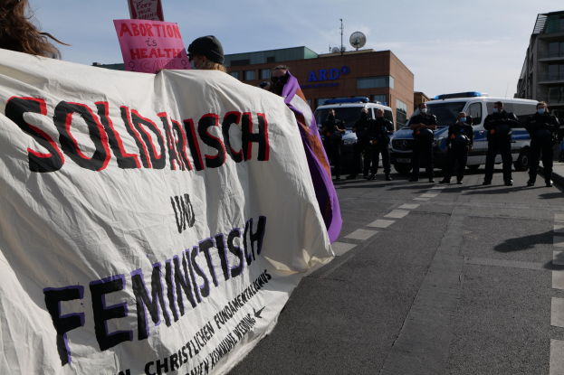 Gruppe von Menschen marschiert auf einer Straße und hält ein 'Solidarität und Feminismus'-Schild, mit geparkten Autos und Gebäuden im Hintergrund unter einem klaren blauen Himmel.