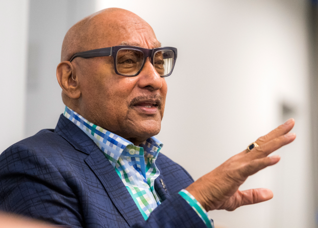 Ein Mann im Anzug und mit Brille spricht in ein Mikrofon und blickt auf ein unscharfes Publikum.