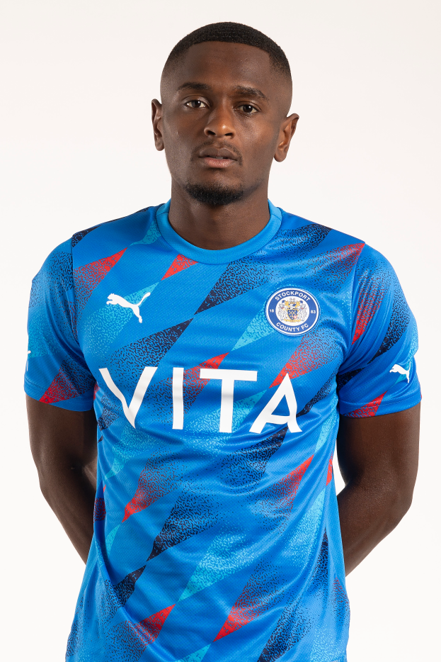 Ein Mann in einem blauen Leicester City Away-Shirt von der Saison 2018-19, das das Team-Logo, einen Rundhalsausschnitt und kurze Ärmel aufweist, steht mit den Händen in den Taschen und einem leichten Lächeln.