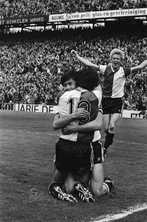 Drei Fußballspieler umarmen sich auf dem Boden nach einem Tor, umgeben von einer jubelnden Menge, die Fahnen schwingt in einem schwarz-weißen Stadion Setting.