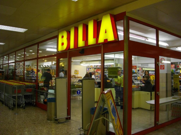Ein Supermarkt mit einem Eingang in der Mitte, Mülltonnen daneben, eine Reihe von Einkaufswagen auf der linken Seite, Deckenbeleuchtung und einige Menschen im Inneren, die Produkte anschauen.