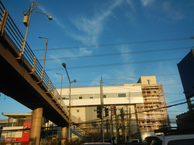 Stadtstraße mit parkenden Autos, eine Brücke mit Geländern, Laternenpfähle, Strommasten mit Drähten, Ampeln, Gebäude mit Fenstern und ein bewölkter Himmel im Hintergrund.