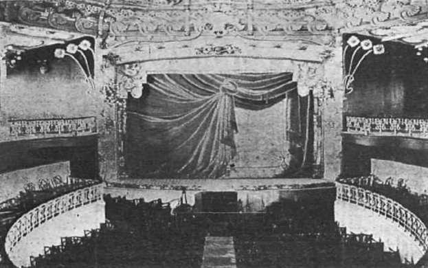 Altes Schwarz-Weiß-Foto des Theatre Royal Auditoriums in London, das eine Bühne mit Vorhang, Geländer und ornamentale Wanddesigns zeigt, mit einigen sitzenden Menschen und Text unten.