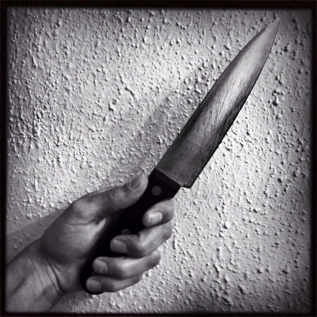Eine Person Hand, die ein Messer hält, mit einer Wand im Hintergrund, auf einem schwarz-weißen bearbeiteten Bild.