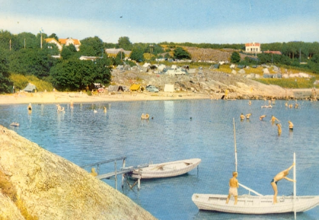 Ein altes Foto von Menschen, die in der Nähe eines Bootes schwimmen, mit einer Brücke mit Geländern im Vordergrund, einem Felsen links und einem Hintergrund mit Bäumen, Gebäuden, Fahrzeugen, Pfählen und einem bewölkten Himmel.