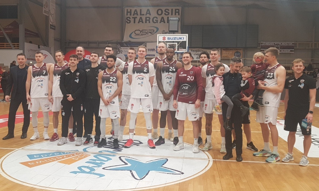 Männer- und Kinder-Gruppe auf einem Basketballfeld mit Schildern, die "Hala Osir Starga" lesen, sitzende Zuschauer, ein Basketballkorb, Metallstangen, Deckenleuchten und eine Wand im Hintergrund.