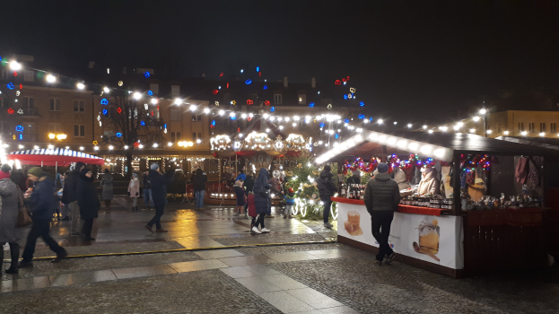 Ein belebter Weihnachtsmarkt bei Nacht mit Menschen, geschmückten Buden, beleuchteten Gebäuden, Bäumen und festlicher Beleuchtung am Himmel.