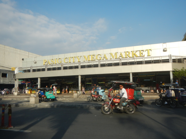 Eine belebte Straßenszene am Pasig City Mega Market mit Fahrzeugen und Fußgängern, mit einem Gebäude mit sichtbarem Text im Hintergrund unter einem bewölkten Himmel.
