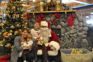 Eine Frau, Kinder und ein Mann in Santa-Klaus-Kost√ľm mit Brille und Handschuhen stehen neben einem geschm√ľckten Weihnachtsbaum, einer Felswand, Spielzeugen, Deko, einem Korb und Hintergrundbeleuchtung, mit einem Jungen, der ein P√§ckchen h√∂lt.