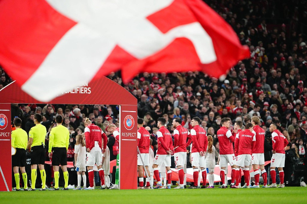 Eine Gruppe von Menschen auf einem Fußballfeld mit einer roten und weißen Flagge im Vordergrund, einem Bogen mit dem Text "Bayern München vs. Bayern München Wetten & Vorschau" im Hintergrund und einer großen Menschenmenge im Stadion.