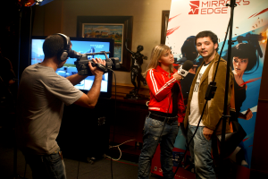 Ein Mann mit einer Videokamera und Headset filmt ein Interview zwischen einem Mann und einer Frau, wobei im Hintergrund ein Fernseher und ein Foto Rahmen zu sehen sind.