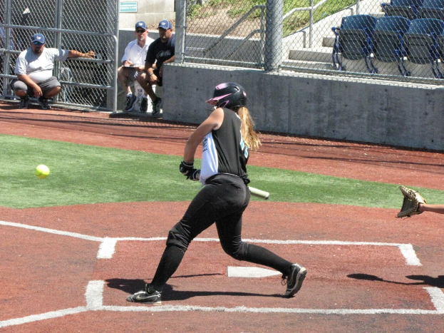 Eine Frau in einem Baseball-Outfit ist bereit, einen Schläger zu schwingen, mit einer Handschuh tragenden Hand hinter ihr, Zuschauern im Hintergrund und einem Zaun mit leeren Stühlen und Gras dahinter.