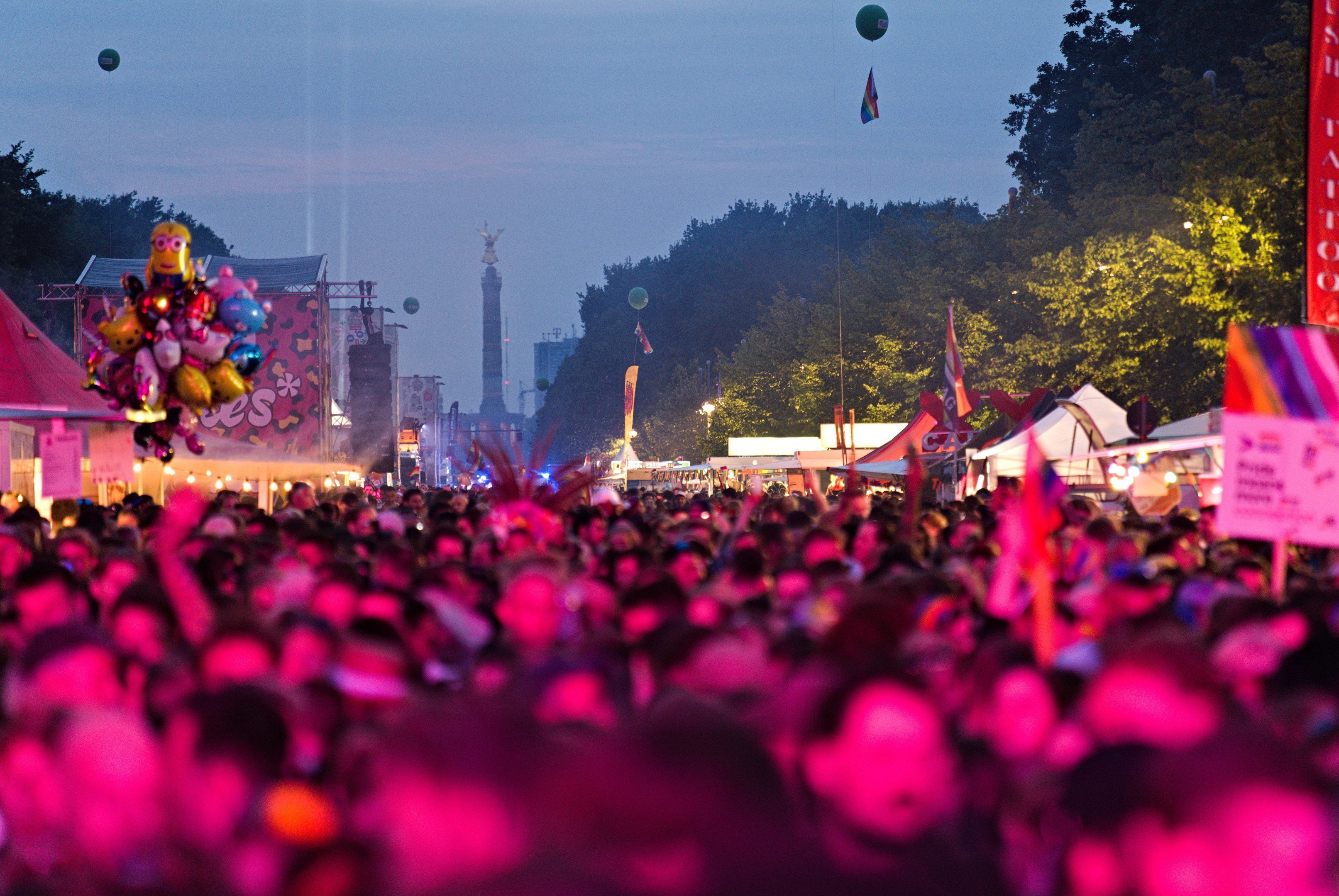 Eine große Menschenmenge auf einem nächtlichen Musikfestival mit bunten Lichtern, Ballons, Zelten, Schildern, Fahnen, Bäumen und einem sternenklaren Himmel im Hintergrund.