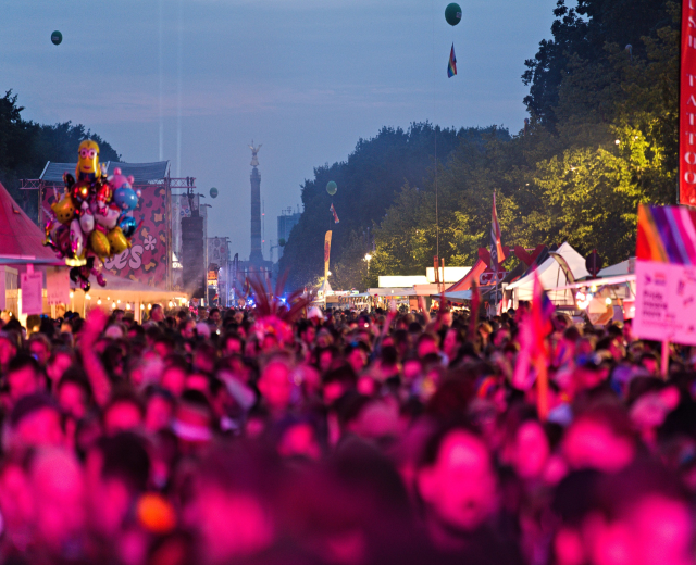 Eine große Menschenmenge auf einem nächtlichen Musikfestival mit bunten Lichtern, Ballons, Zelten, Schildern, Fahnen, Bäumen und einem sternenklaren Himmel im Hintergrund.