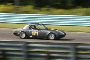 Ein Auto mit einem Helmträger fährt auf einer Straße, die von Barrieren gesäumt ist, mit Bäumen im Hintergrund.