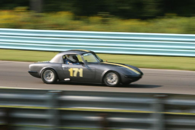 Ein Auto mit einem Helmträger fährt auf einer Straße, die von Barrieren gesäumt ist, mit Bäumen im Hintergrund.