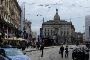 Lebendige Stadtstraße mit einem parkenden Polizeiwagen, Fußgänger mit Taschen, fahrende Fahrzeuge, Gebäude mit Fenstern und Bannern, Laternen, Ampeln und ein bewölkter Himmel.