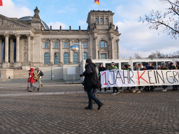 Eine Gruppe von Menschen geht vor dem Reichstaggebäude in Berlin vorbei, eine Fahne haltend, mit dem Gebäude, das Säulen, Fenster, Bögen, Statuen und Flaggen hat, umgeben von einem Metallzaun, einem Straßenpfahl, einer Straßenlaterne, Bäumen und einem bewölkten Himmel im Hintergrund.