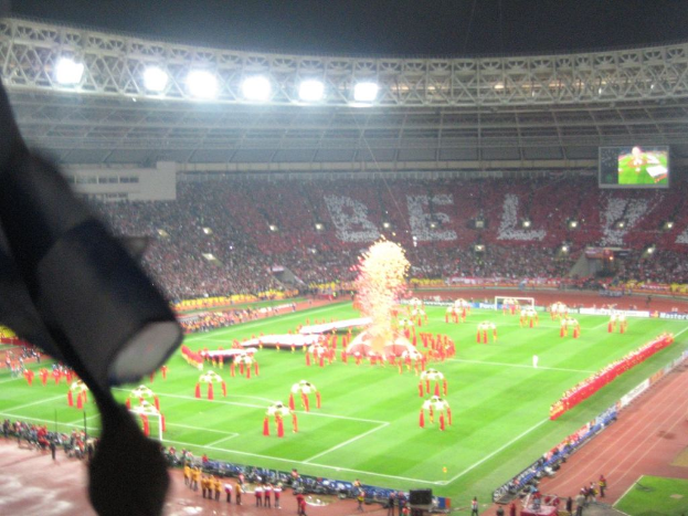 Ein Stadion mit Flutlicht, Eisengittern, Zuschauern, stehenden Personen, Werbetafeln, Anzeigetafeln, Überwachungskameras, Fahnen und Dekorationen.