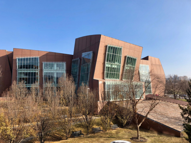 Großes Gebäude mit vielen Fenstern, umgeben von Bäumen, Bänken, Gras und einem Laternenmast, mit anderen Gebäuden und einem klaren blauen Himmel im Hintergrund; als der neue Campus der University of Illinois at Chicago identifiziert.