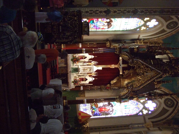 Eine Innenansicht einer Kirche mit einem Kreuz, das eine Jesus-Skulptur zeigt, und zahlreichen Menschen, die davor auf Stühlen sitzen.